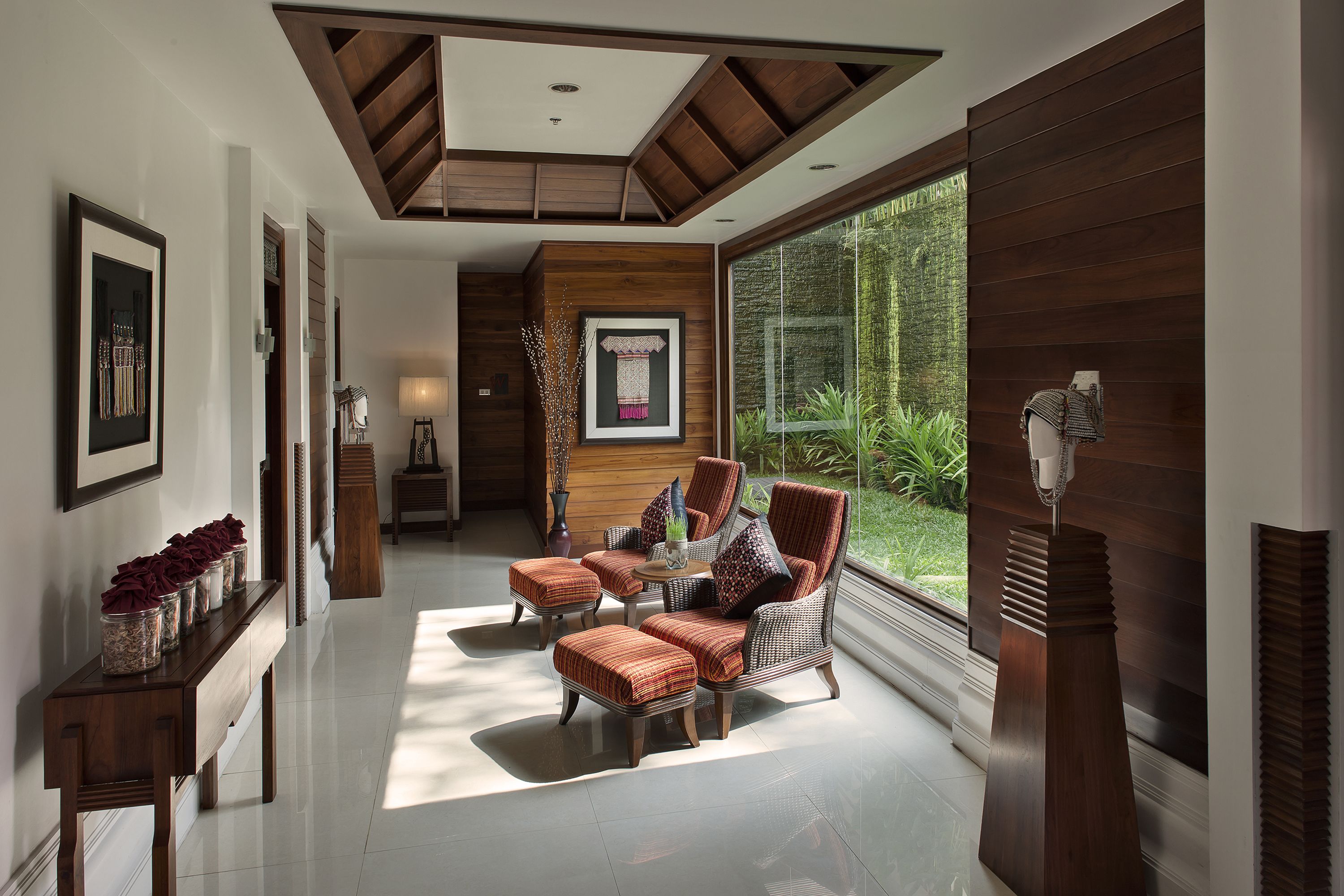 Um interior moderno com teto de madeira, uma janela grande com vista para o verde, uma área de estar com mesa e um console com objetos decorativos.