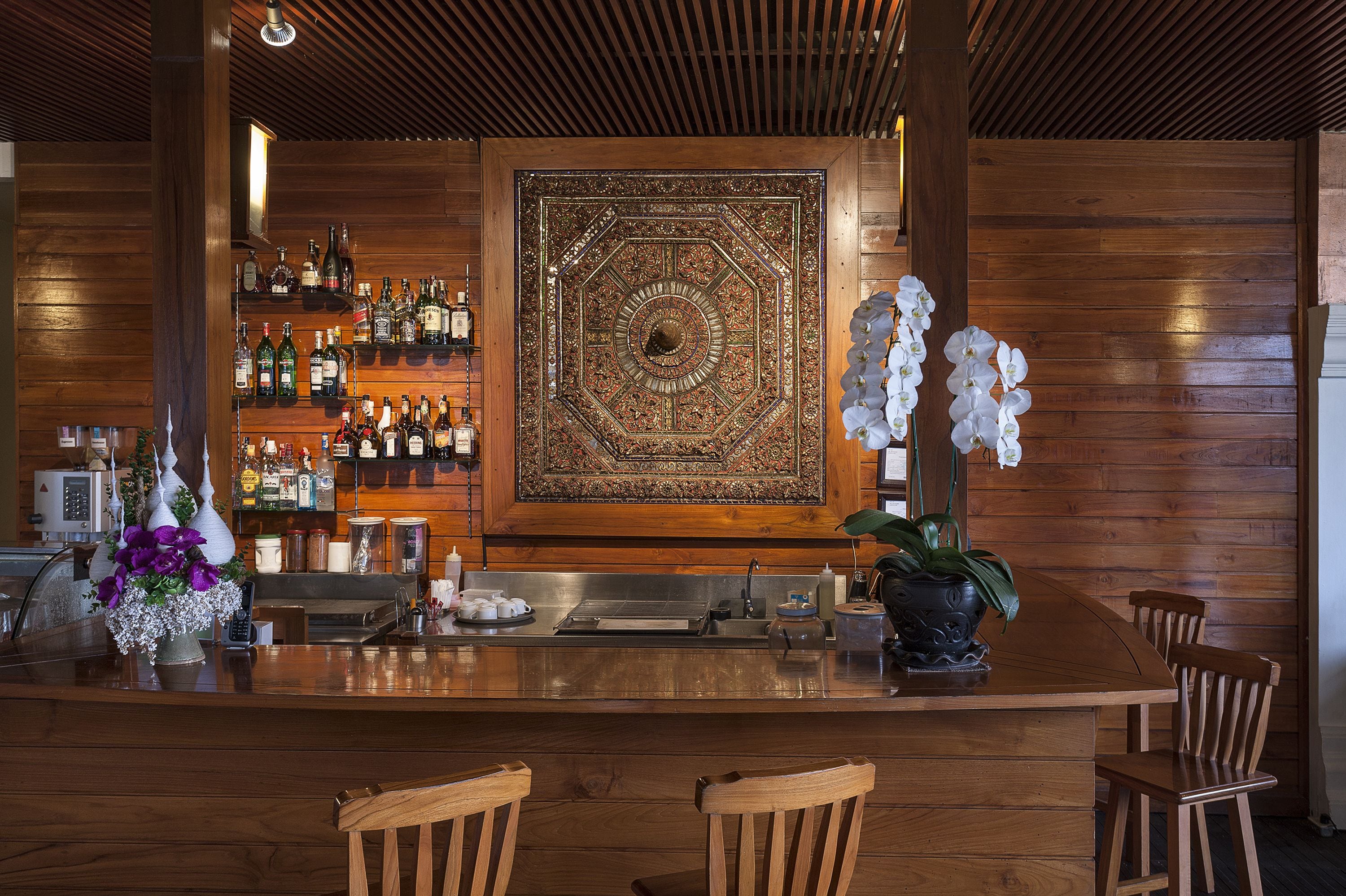 A wooden bar area with a large decorative wall panel, shelves of liquor bottles, and two chairs.