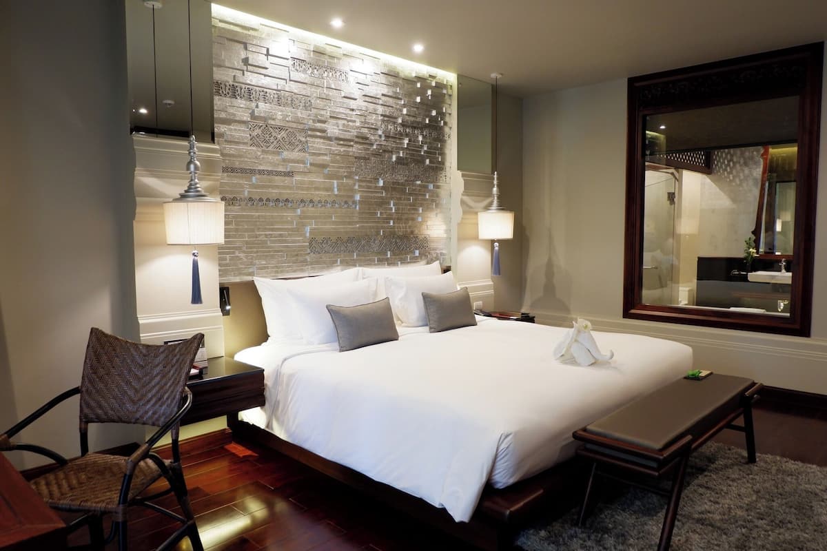 Quarto de hotel com uma cama grande, cabeceira de madeira, mesa de cabeceira, uma cadeira, escrivaninha e um espelho amplo.