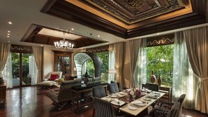 Une salle à manger spacieuse avec un plafond haut, de magnifiques lustres et de grandes fenêtres offrant une vue sur une végétation luxuriante.