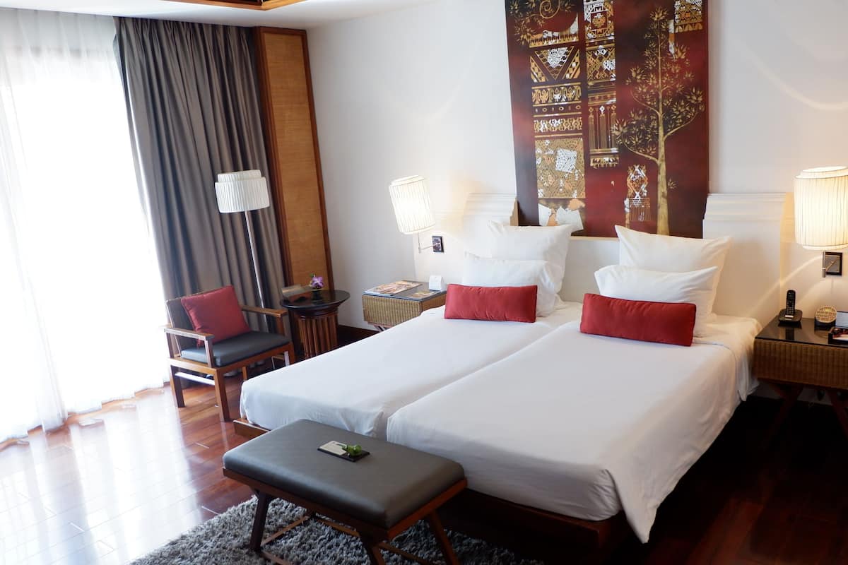Quarto de hotel com duas camas, uma escrivaninha, uma cadeira, um abajur e um grande tapeçário na parede.