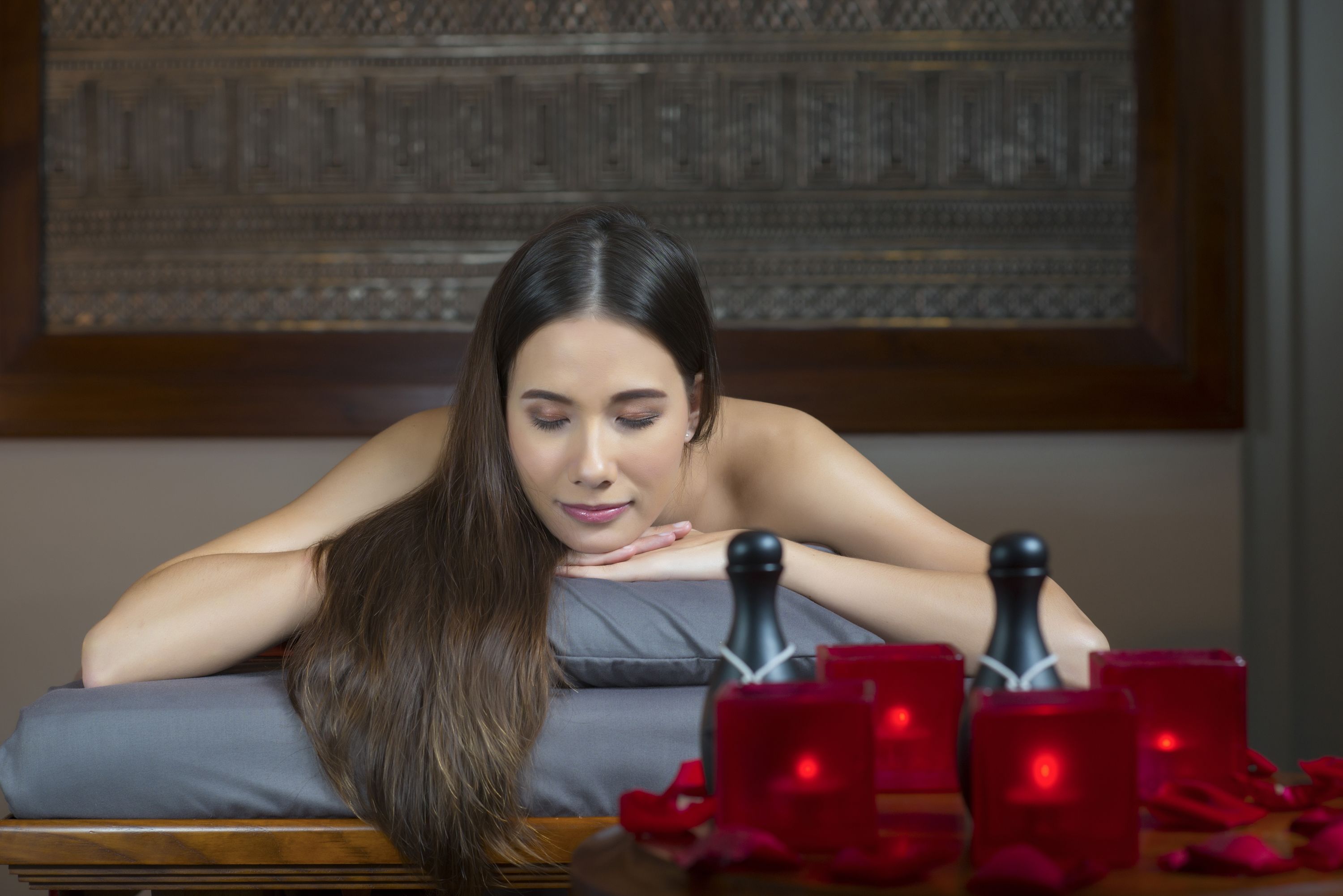A person reclining on a bed with a pillow, surrounded by red candles and rose petals.