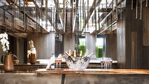 A bar area with a wooden counter, stools, a decorative ice bucket with glasses, and a large window in the background. - Jiva Hill Resort - Genève (Crozet)