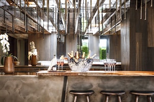 Uma área de bar com balcão de madeira, banquetas, um balde de gelo decorativo com copos e uma janela grande ao fundo.