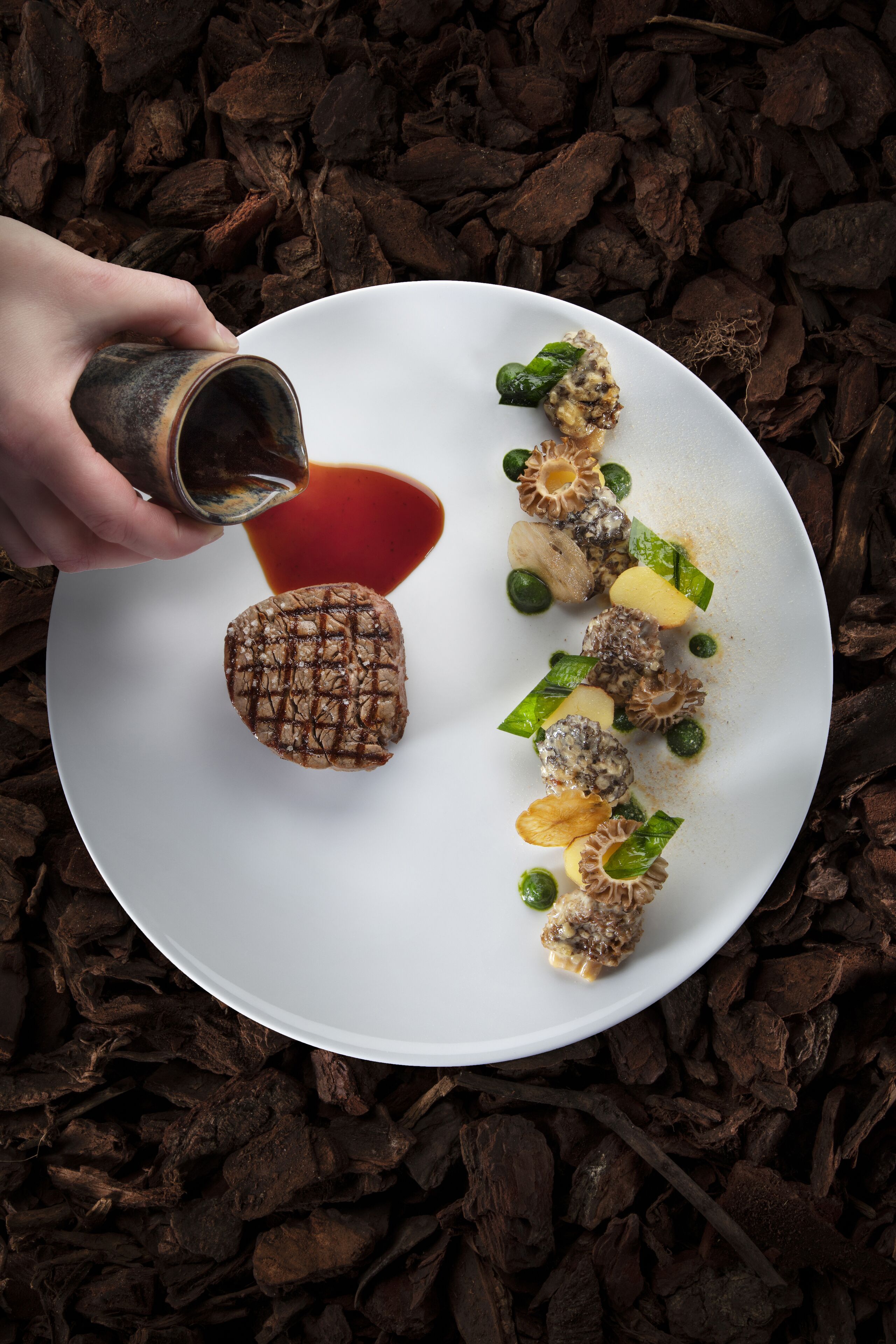 A white plate with a grilled steak, green vegetables, and mushrooms, with a hand pouring sauce.