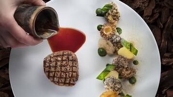 A white plate with a grilled steak, green vegetables, and mushrooms, with a hand pouring sauce.