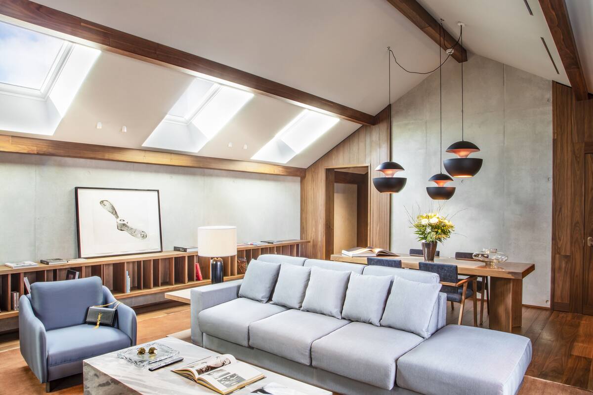 A modern living room with a sofa, armchair, and coffee table. There is a dining area with a table and chairs. The room has wooden beams on the ceiling and a skylight.