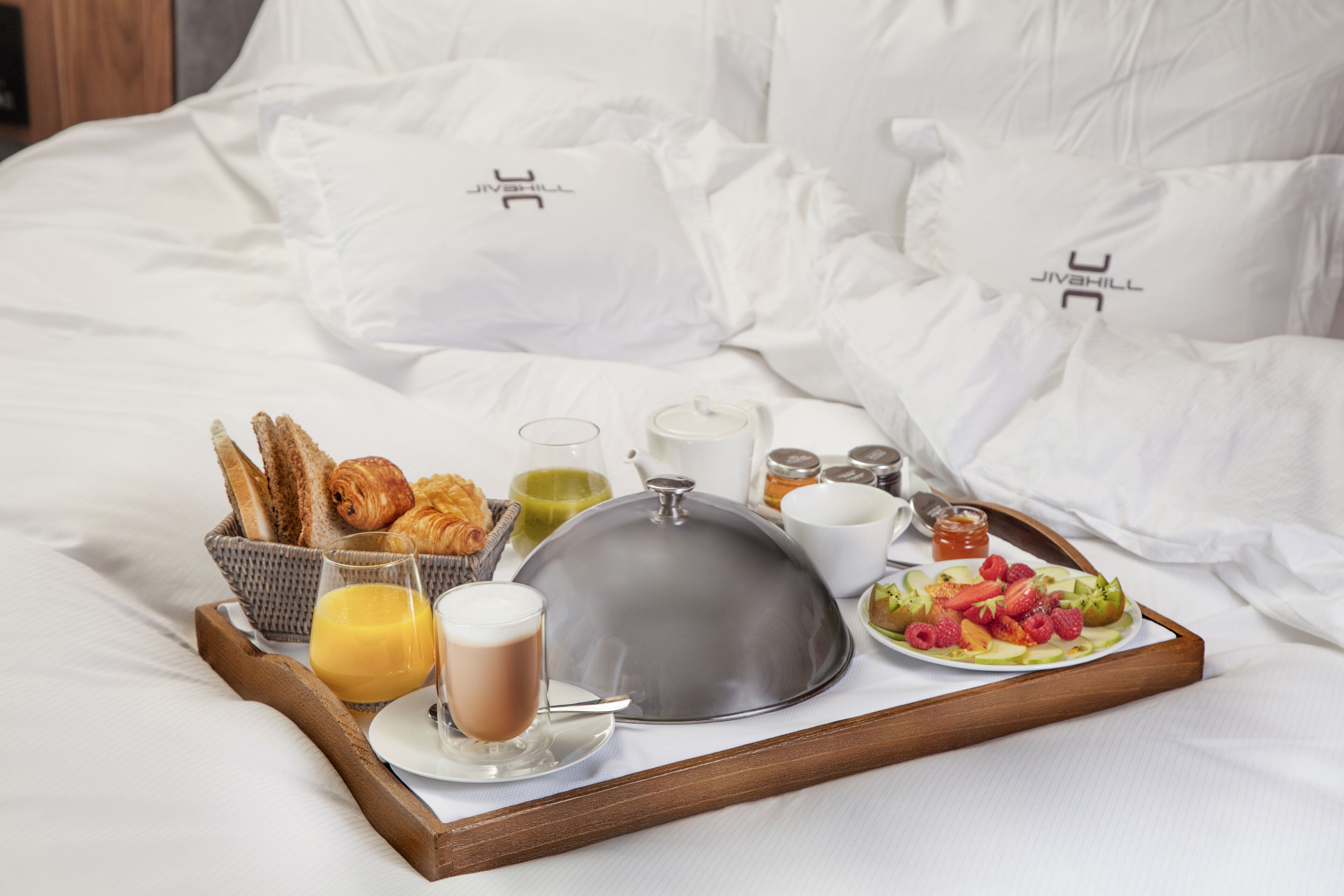 Bandeja de desayuno con croissants, fruta y bebidas sobre una cama con ropa blanca y almohadas.