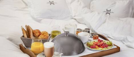 Bandeja de desayuno con croissants, fruta y bebidas sobre una cama con ropa blanca y almohadas.