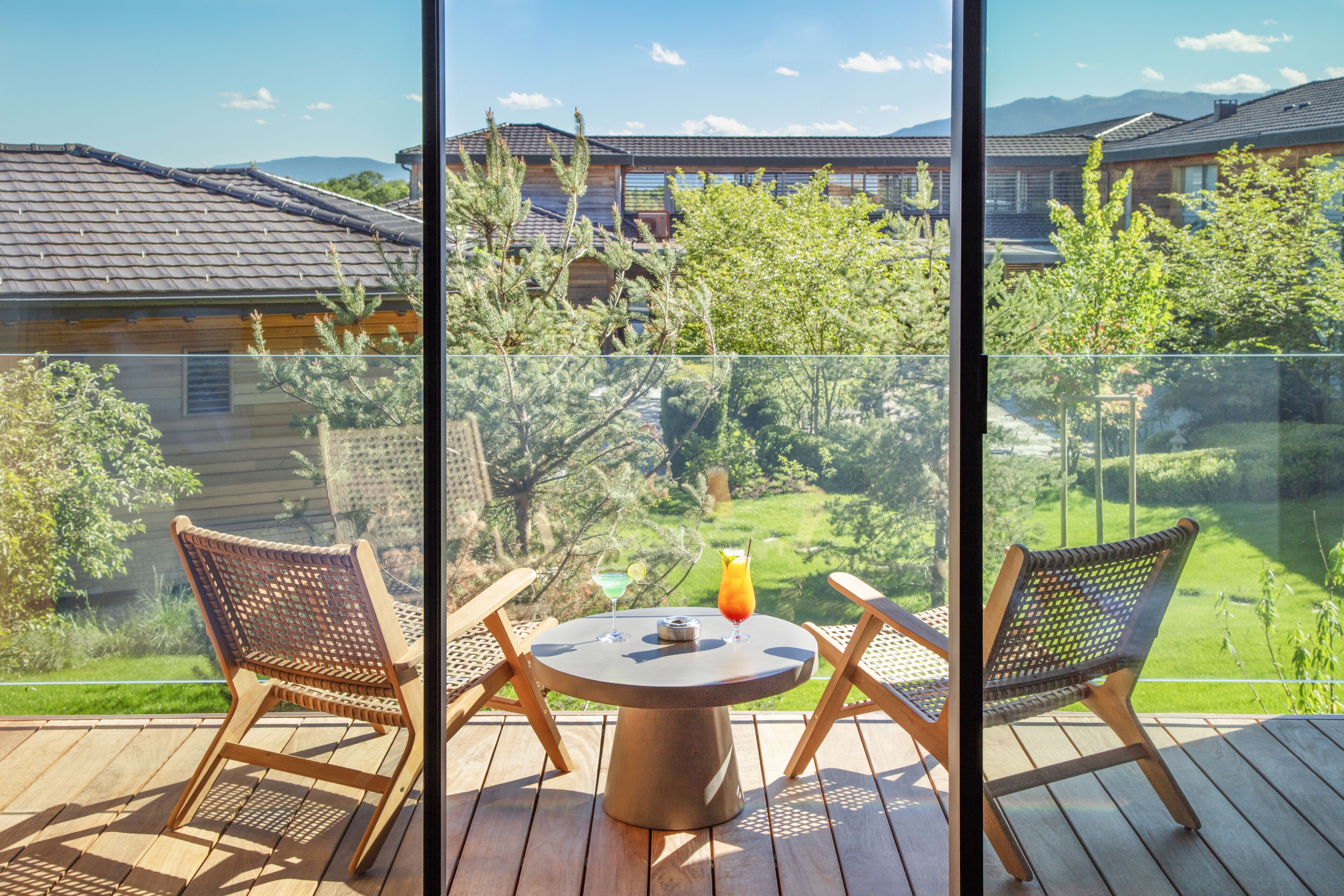 Een balkon met een ronde tafel, twee rieten stoelen en een glazen reling.