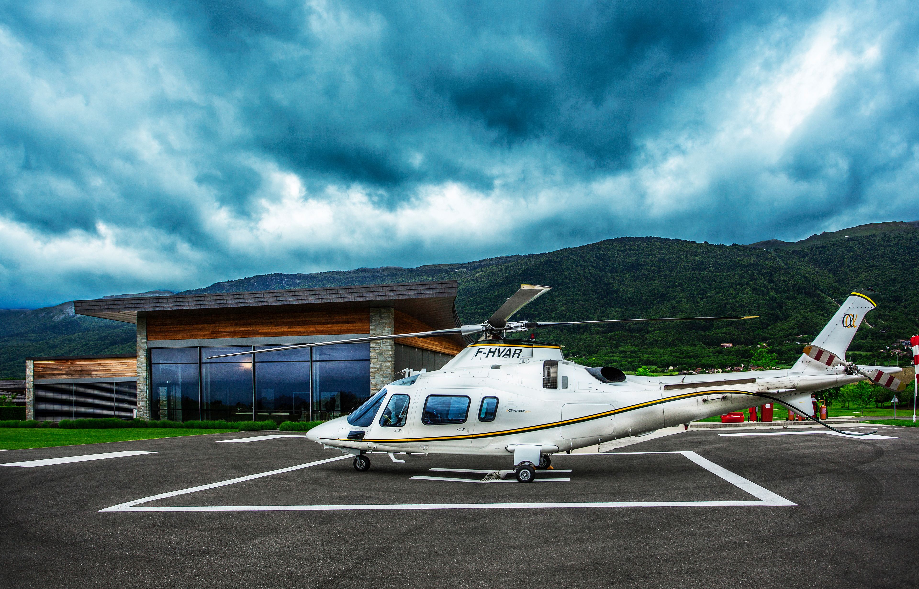 Een witte helikopter met 'F-HVAR' op de staart geparkeerd op een tarmac, met een gebouw en bergen op de achtergrond.