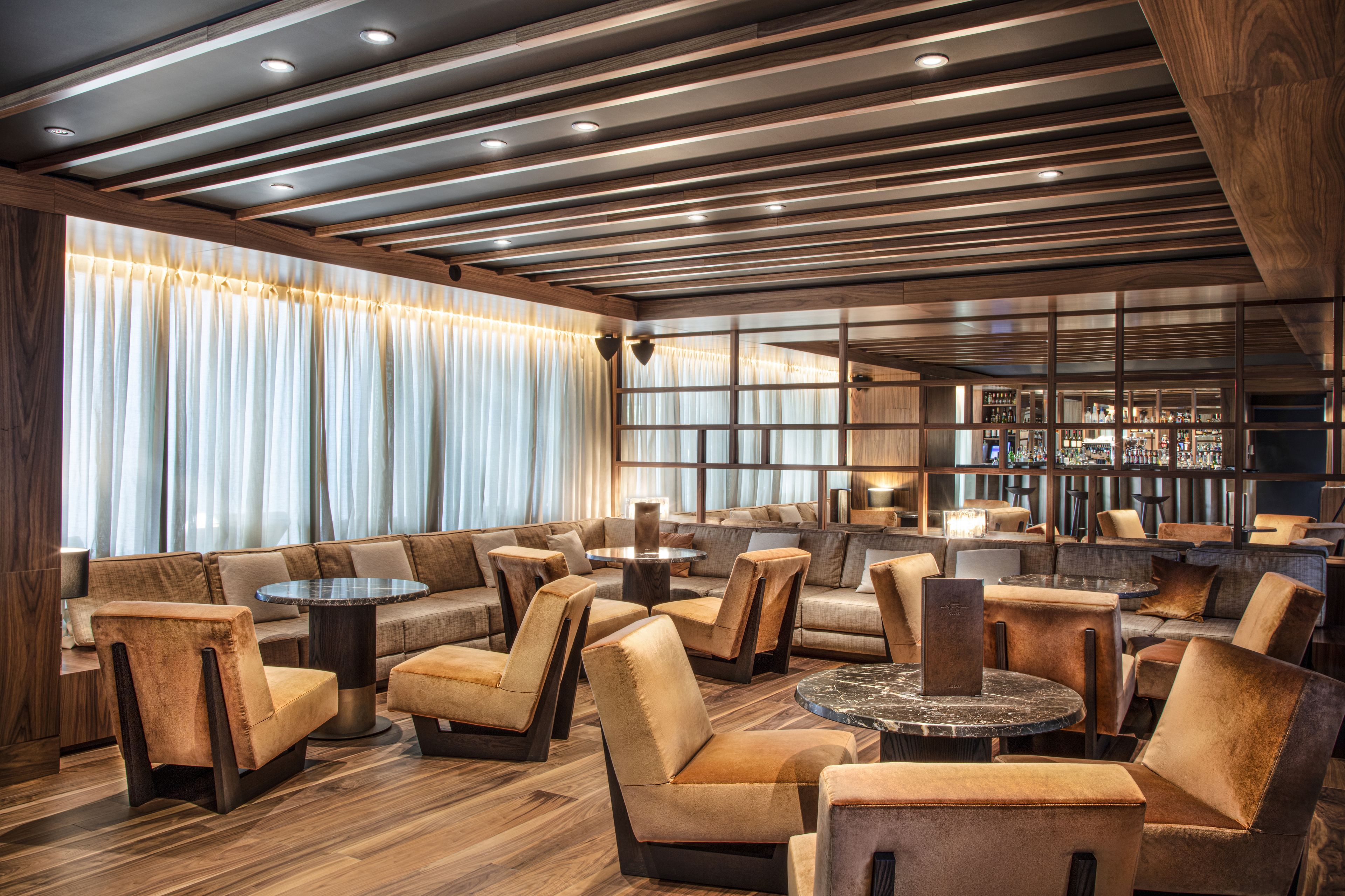 Un espace de détente moderne, doté de canapés, de fauteuils et de tables, avec un plafond en bois et de grandes fenêtres agrémentées de rideaux.
