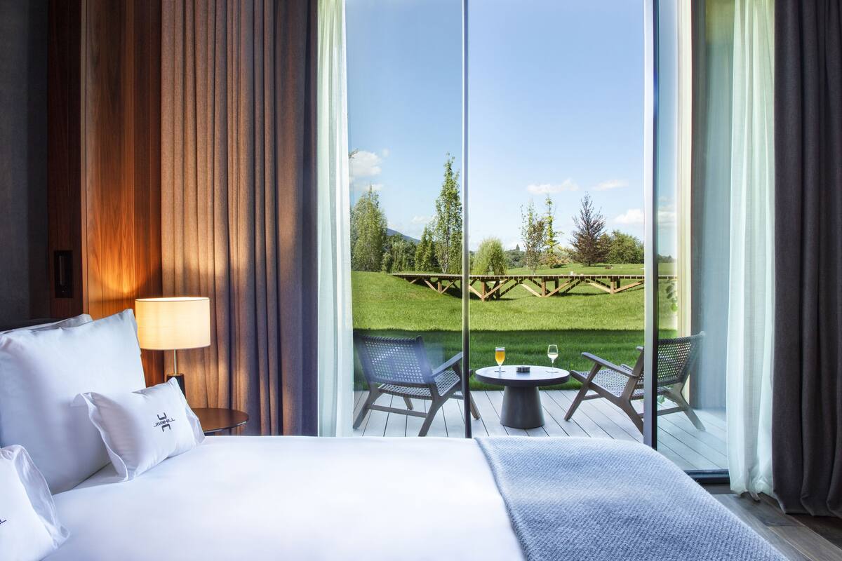 A hotel room with a large bed, a bedside table with a lamp, and a view of a grassy outdoor area with a wooden deck.