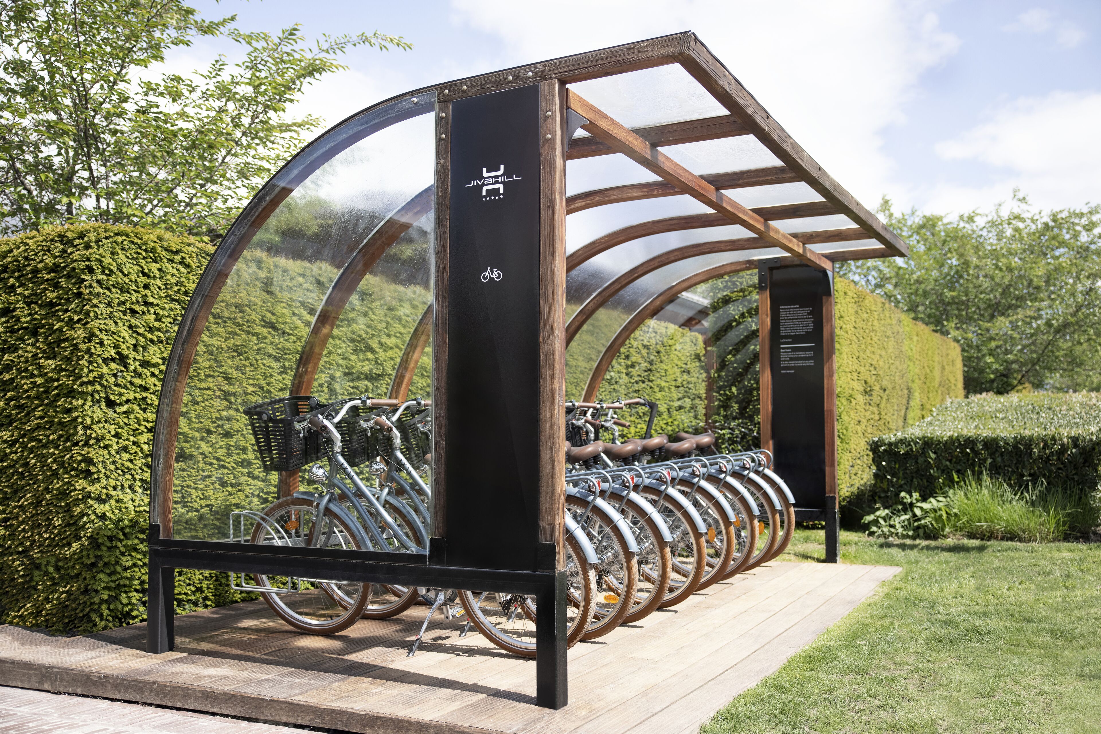 A bike rental station with a transparent roof and wooden structure, surrounded by greenery.