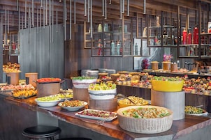 Buffet com diversos alimentos e bebidas, incluindo saladas, frutas e doces, expostos em aparadores e mesas de madeira.