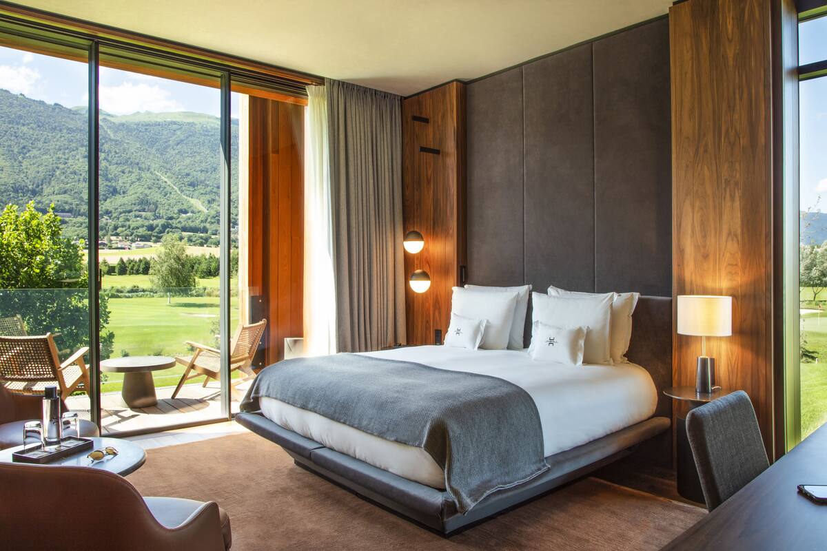A spacious bedroom with a large bed, a view of a lush green landscape, and a balcony with seating.