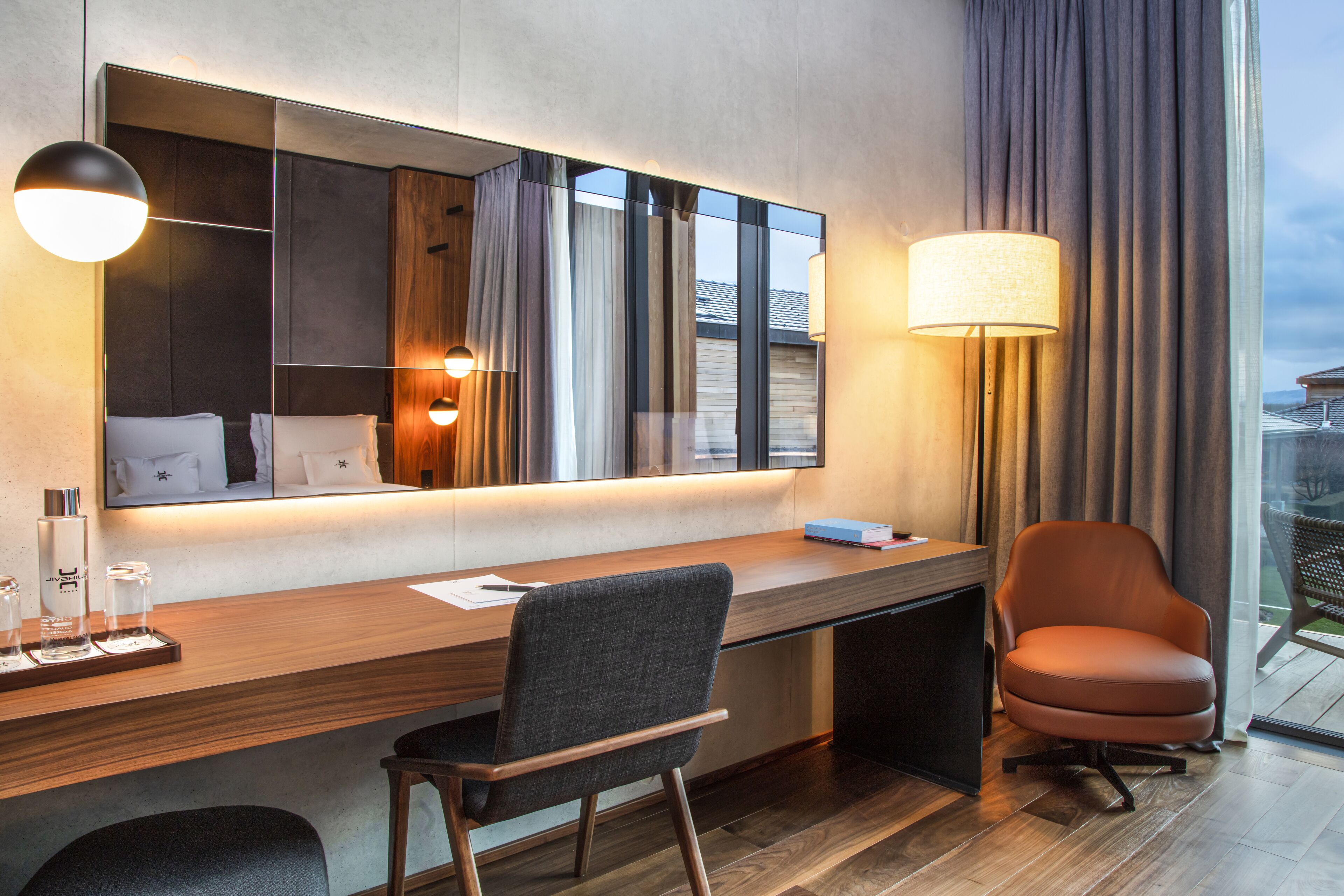 A modern hotel room with a wooden desk, a comfortable chair, a large mirror, and a view of the outdoors.