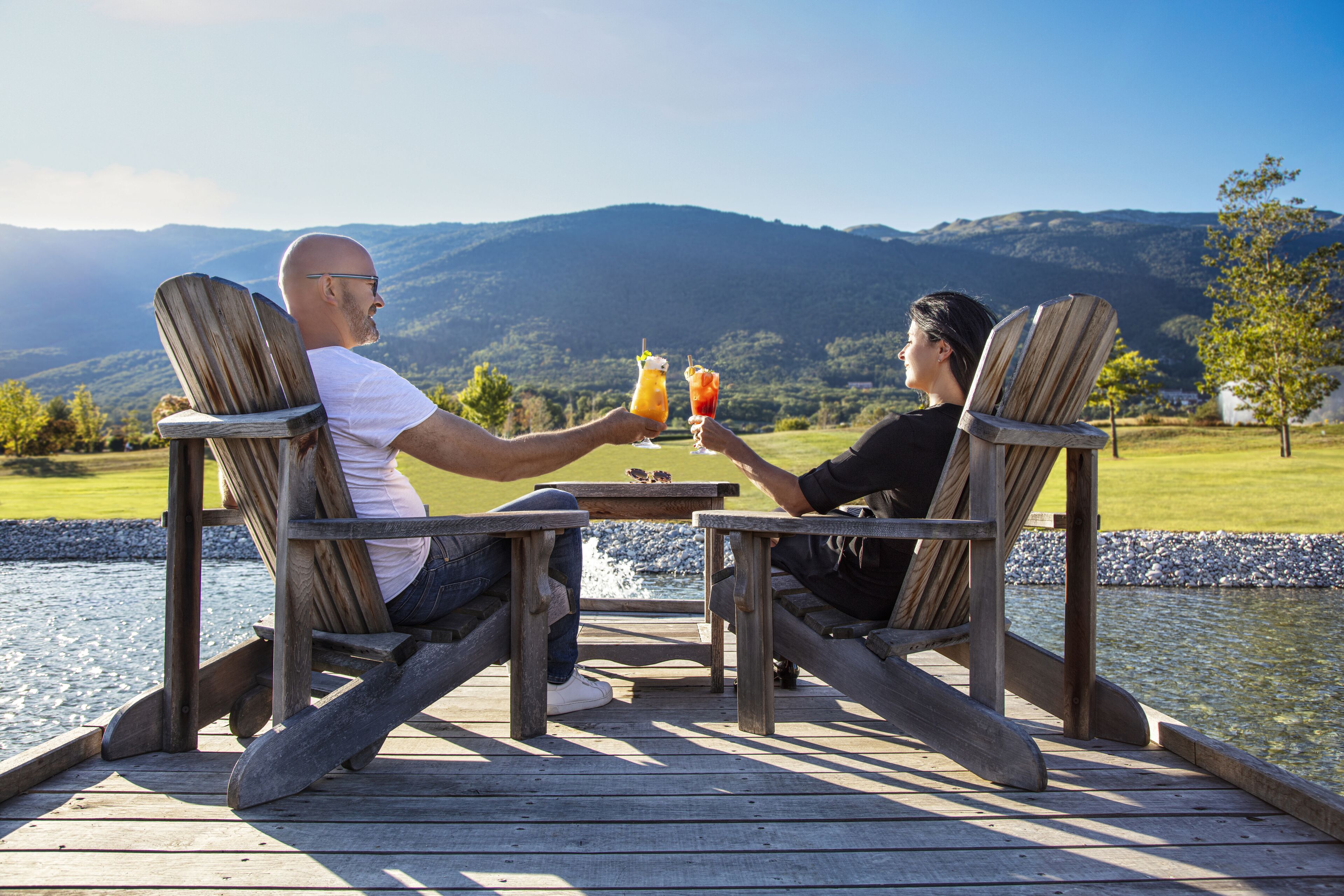 Two people seated at a wooden table on a deck overlooking a body of water and mountains.