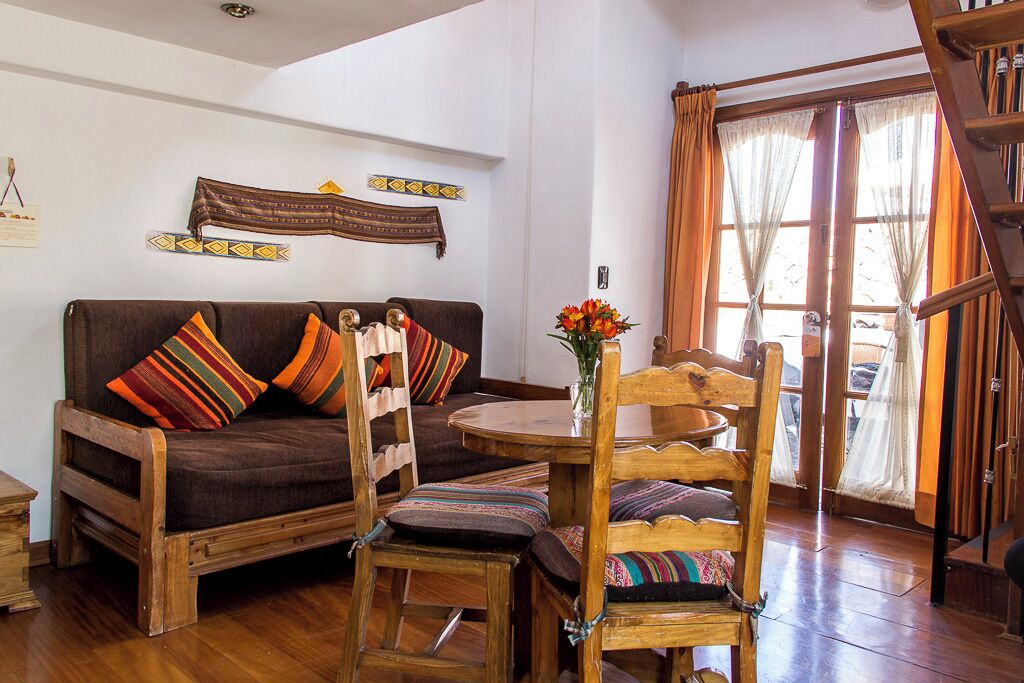 A cozy living room with a brown sofa, wooden chairs, and a small table. There are colorful cushions on the sofa and chairs. A wooden ladder is visible in the background.