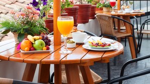 A wooden table with a fruit platter, a glass of orange juice, and a plate of food, set under a large umbrella on a terrace with potted plants and a view of a tiled roof. - Casa San Blas Boutique Hotel (Cusco)