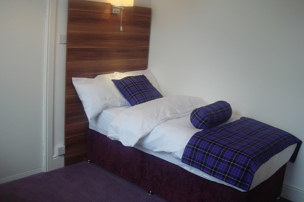 Ein kleines Bett mit Holz-Kopfstück, weißer Bettwäsche und einer violetten Decke.