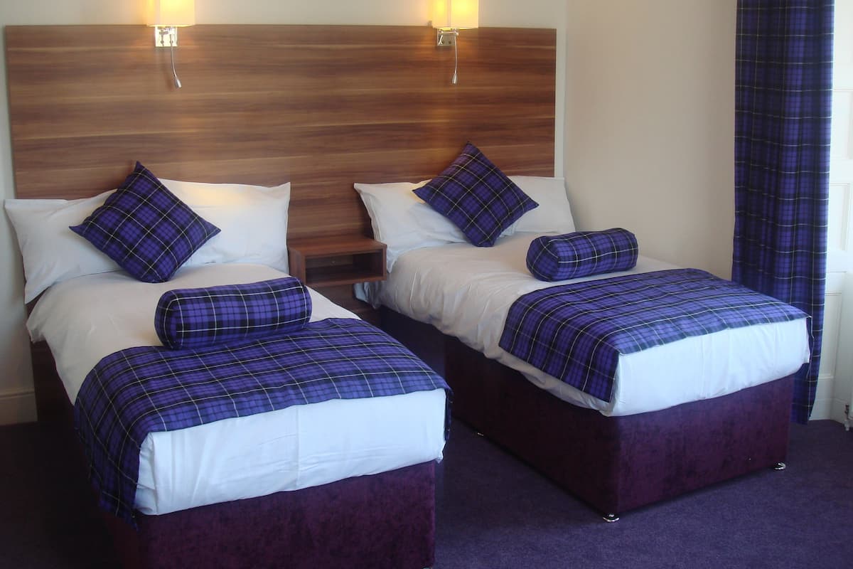 Ein Hotelzimmer mit zwei Betten, jeweils mit einer violett-karierten Tagesdecke und weißen Kissen. An beiden Seiten des Kopfteils befinden sich jeweils eine Wandlampe.
