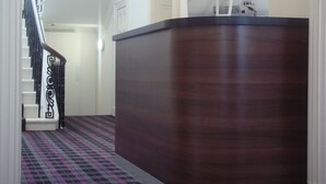 A hallway with a dark wooden reception desk, a patterned carpet, and a staircase with black railings. - Scotia Airport Hotel (Paisley)