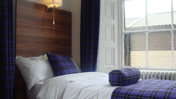 A neatly made bed with white and blue bedding, a wall-mounted lamp, and a window with a view of a brick building. - Scotia Airport Hotel (Paisley)