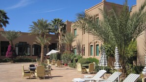 Une cour aménagée avec des fauteuils de jardin, des tables et des parasols, entourée de palmiers et bordée d’un bâtiment à la façade beige.