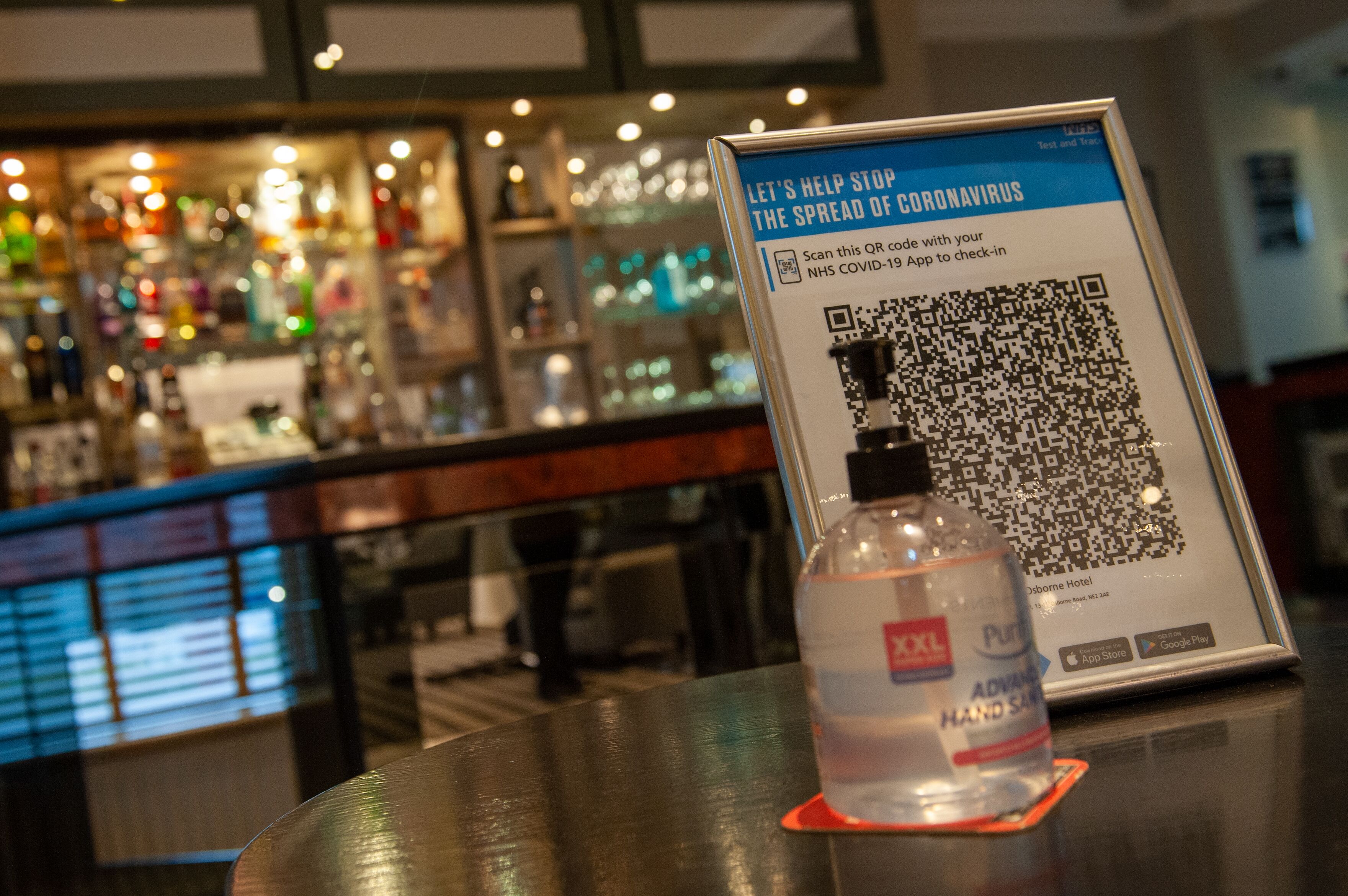A bar area with a QR code for COVID-19 check-in on a table, a bottle of hand sanitizer, and a framed sign.