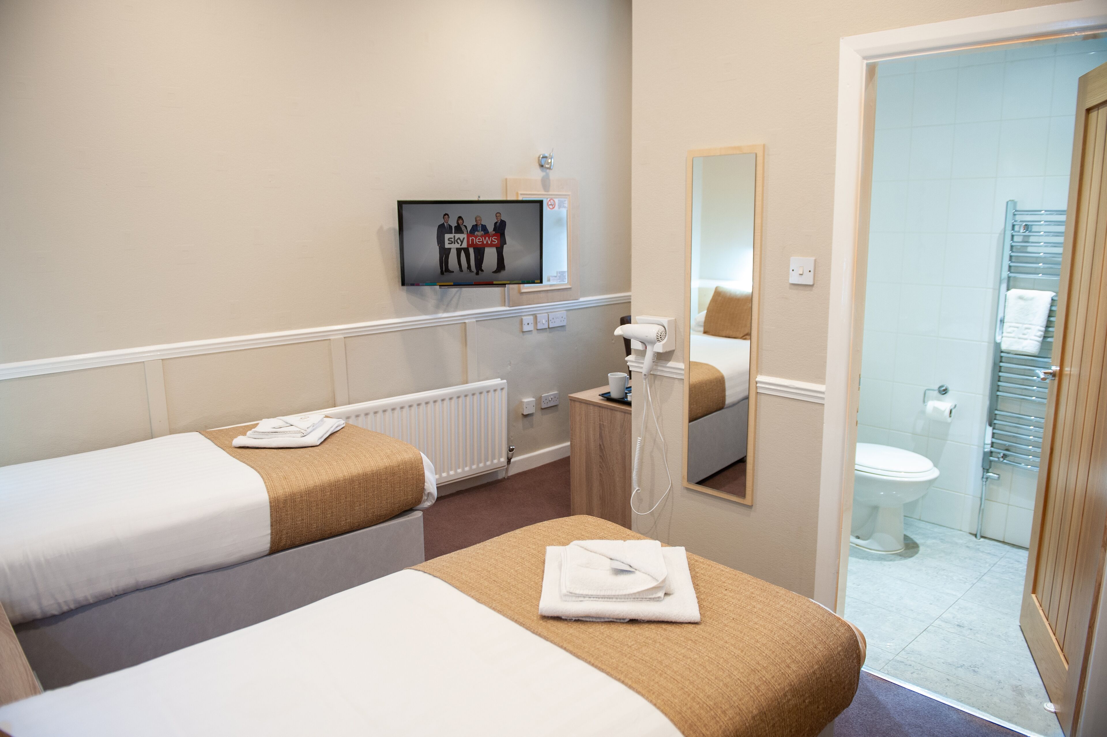 Een hotelkamer met twee bedden, een televisietoestel waarop een nieuwskanaal draait, een badkamer met toilet en handdoekhouder, en een spiegel.