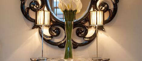 Ein Spiegel mit aufwendigem schwarzem Rahmen, eine Vase mit weißen Blumen und zwei Wandleuchten auf einer Marmorkonsole.