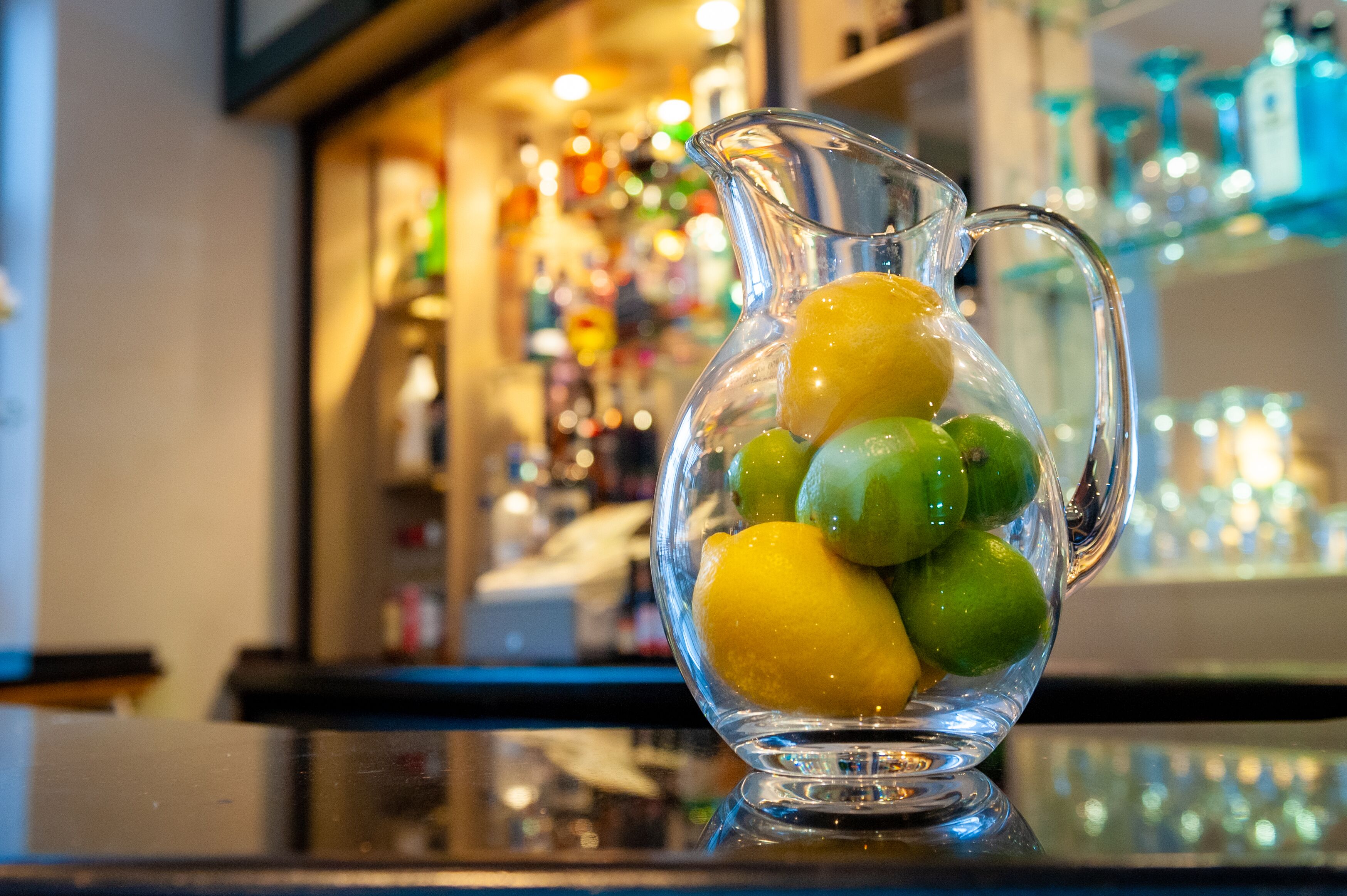 A clear glass pitcher filled with lemons and limes on a reflective surface.