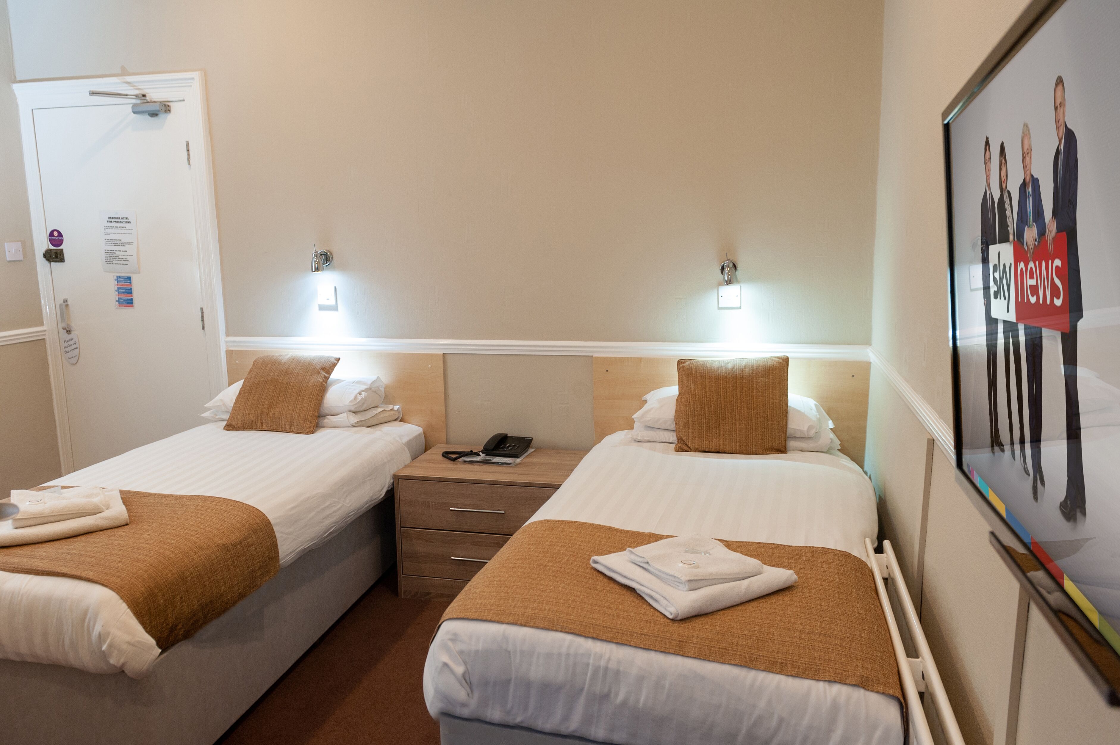 A hotel room with two beds, each with a brown throw blanket and white linens. There is a nightstand with a phone and a small table lamp between the beds. The room has a white door with a sign above it and a framed picture on the wall.