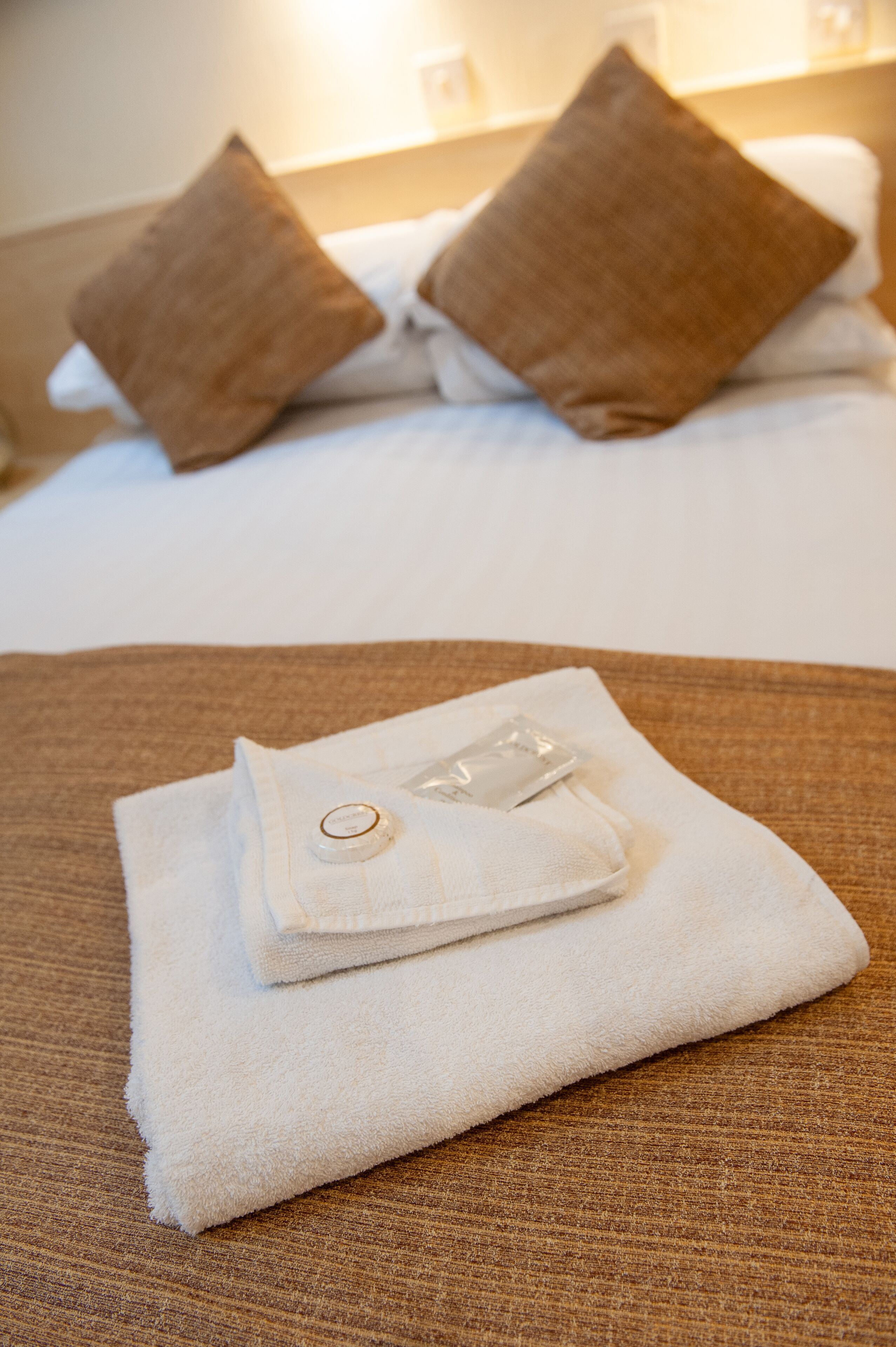 A neatly folded white towel with a small bottle on a brown textured surface, likely a bed or couch, in a hotel room with two brown pillows and a white bedspread.