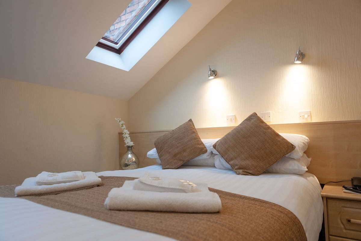 A hotel room with a skylight, a bed with white linens and brown pillows, a bedside table with a vase of flowers, and a nightstand with a lamp.