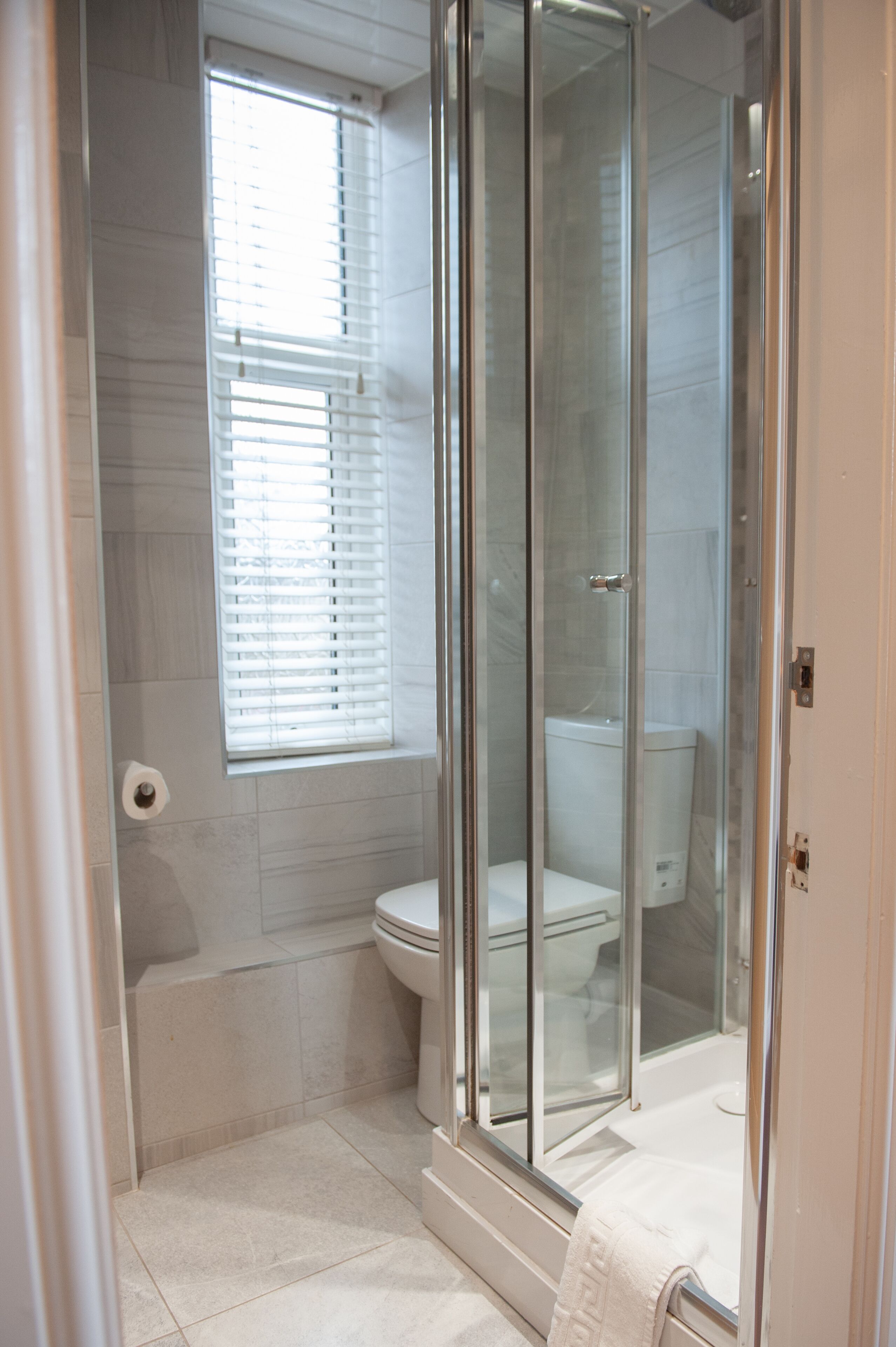 A bathroom with a glass-enclosed shower, a toilet, and a window with blinds.