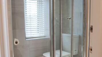 Een badkamer met een douchecabine van glas, een toilet en een raam met zonwering.