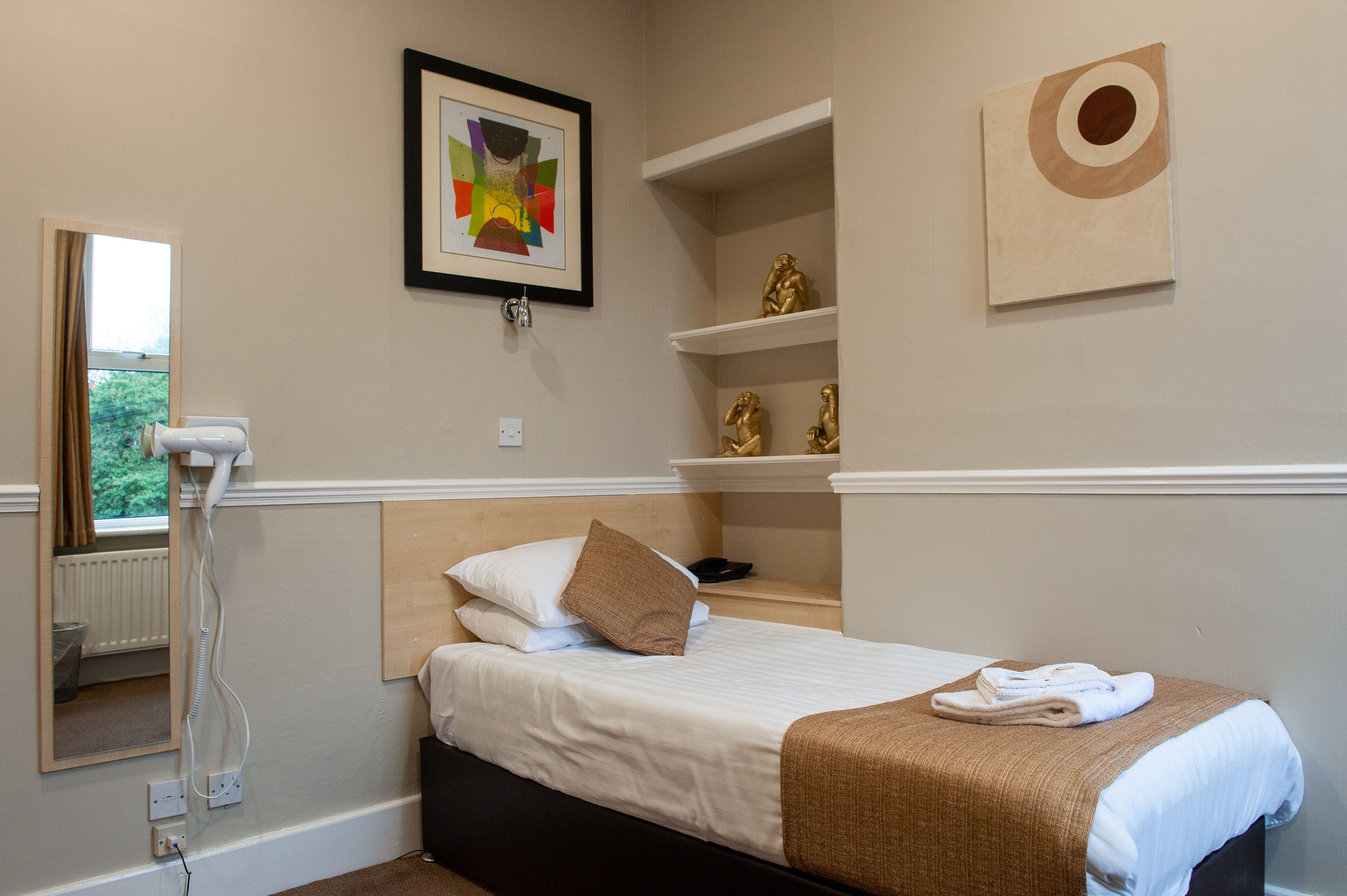 Una habitación de hotel con cama, espejo, secador de pelo, una obra de arte enmarcada y una estantería con objetos decorativos.