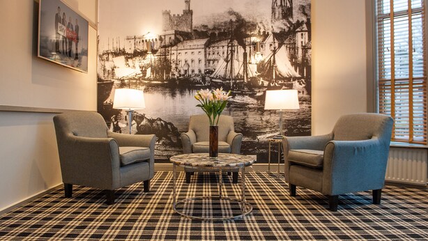 A lobby with two armchairs, a round table with a vase of flowers, and a large black-and-white mural of a harbor scene.