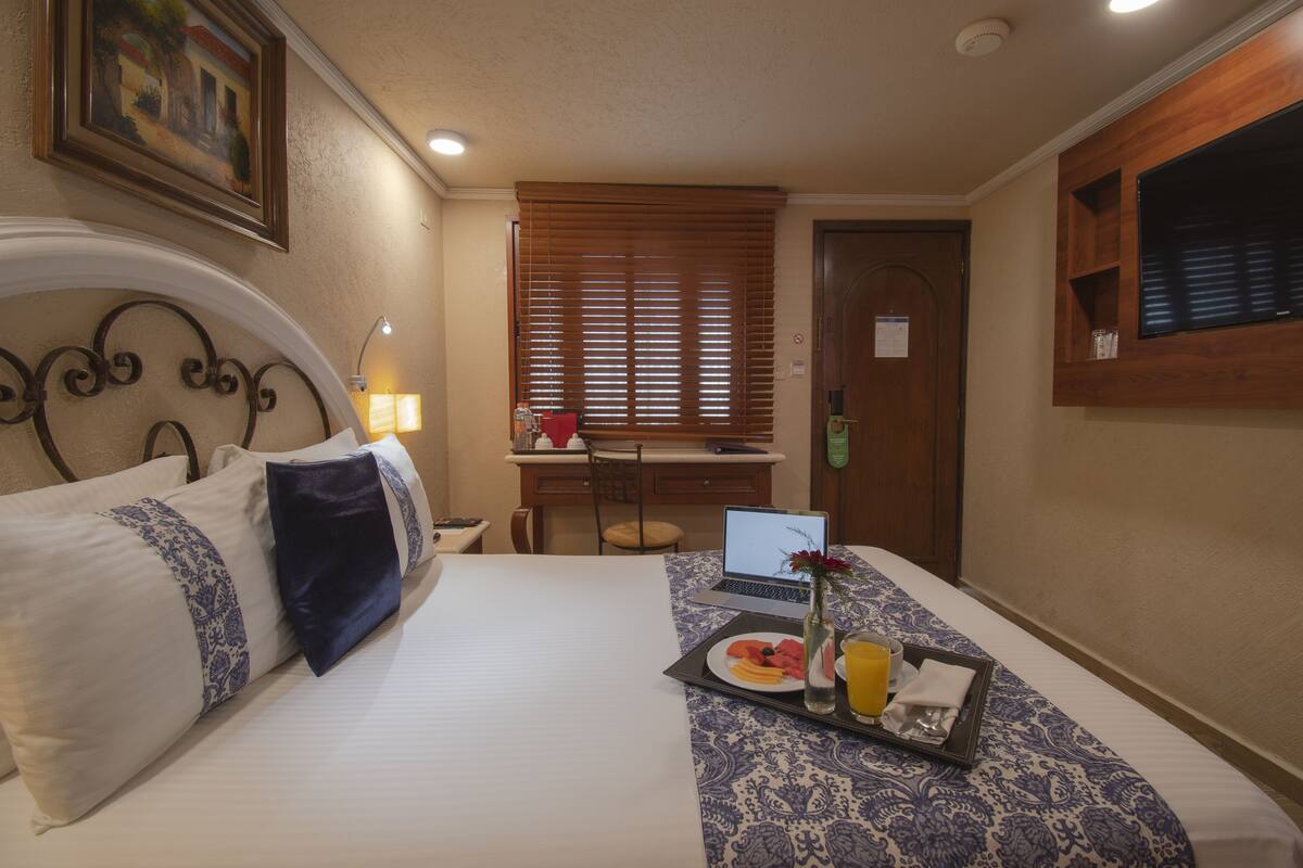 Una habitación de hotel con cama, escritorio con computadora portátil, bandeja de desayuno y ventana con persianas.