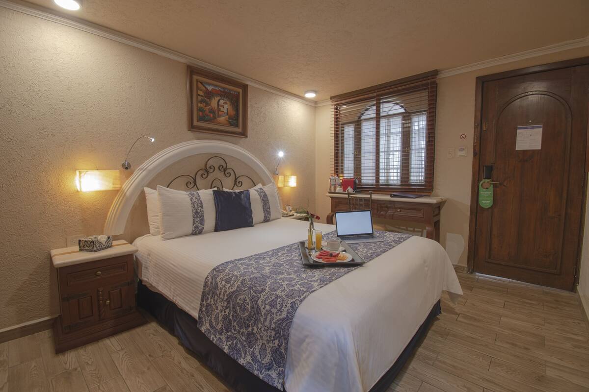 Habitación de hotel con cama, mesita de noche, lámpara, computadora portátil, bandeja con desayuno y una puerta con un letrero verde.