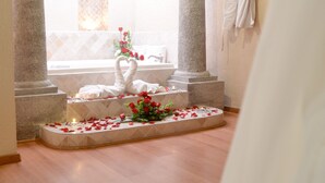 Una habitación con un columnario de piedra, un ventanal con un diseño de abanico decorativo, una bañera y una escalera adornada con pétalos de rosa.