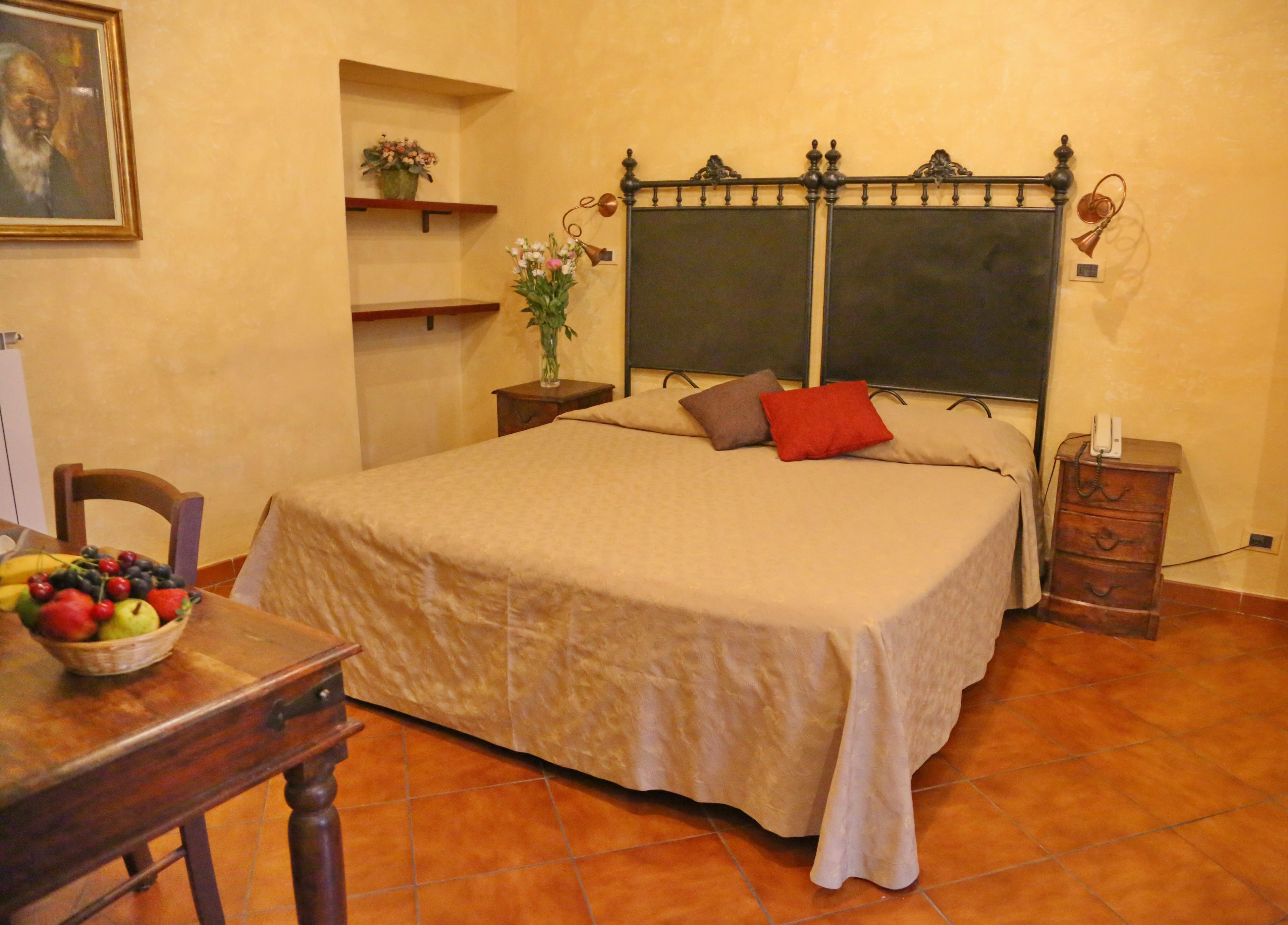 Un dormitorio ordenado con cama, mesitas de noche, una silla, una pequeña mesa con fruta y un estante en la pared con flores.