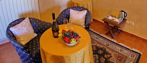 Um quarto acolhedor com uma mesa redonda posta com uma garrafa, copos e uma cesta de frutas. Há duas cadeiras com almofadas, uma mesinha com um vaso de flores e um radiador na parede.