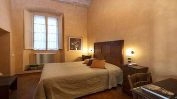 A bedroom with a bed, bedside lamps, a nightstand, a chair, a table, a radiator, a window, and a painting on the wall.