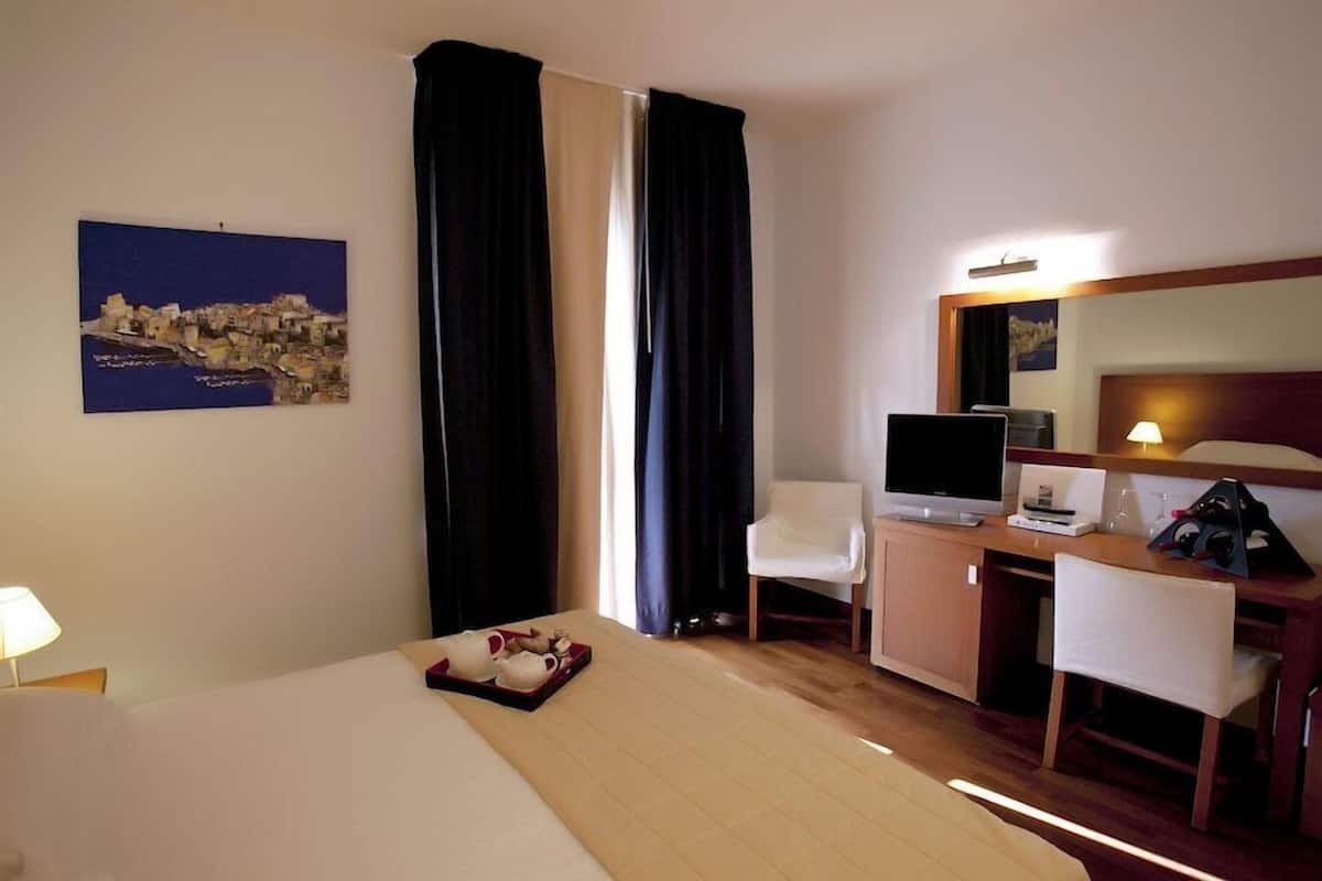 A hotel room with a bed, a desk with a computer, a TV, a chair, a lamp, and a painting on the wall.