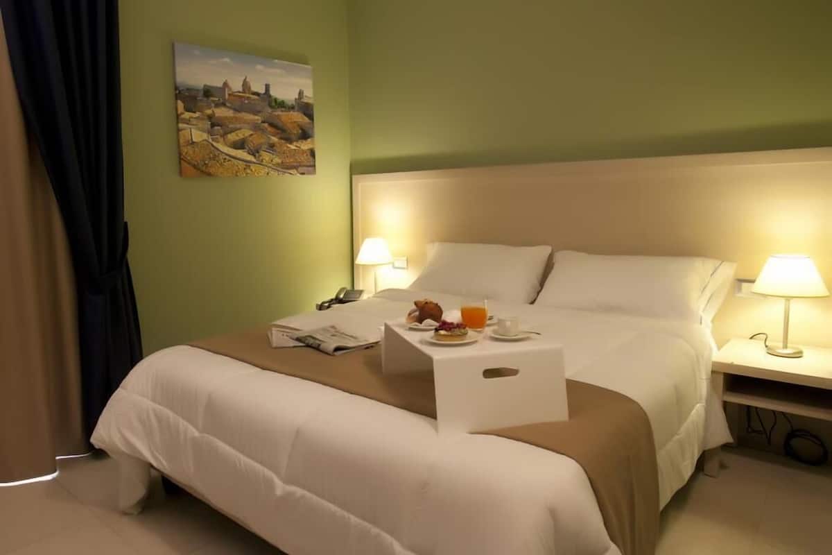 A hotel room with a bed, bedside lamps, a small table with a newspaper, a glass of orange juice, and a painting on the wall.