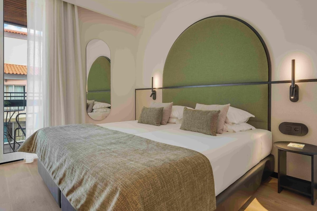 A modern hotel room with a large bed, a round mirror, and a view of the outside balcony.