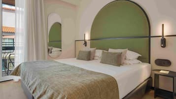 A modern hotel room with a large bed, a round mirror, and a view of the outside balcony.